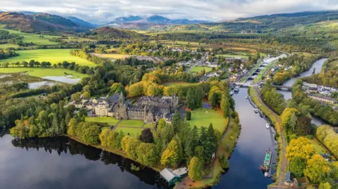 An aerial view over the picturesque village of Fort Augustus on the shores of Loch Ness. The village's abbey is in the foreground next to the loch and a section of the Caledonian Canal, with its locks. There are lots of trees dotted through the village and, in the distance, hills and mountains.