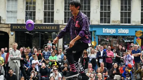 A man wearing a purple pattered adidas jacket riding a unicycle while performing a trick. He has his mouth wide open in an exasperated way. He is standing on front of a crowd of around 50 people many look shocked, happy or bored.