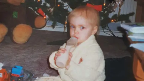 A two-and-a-half year old girl sat on brown carpet. She is wearing a cream fluffy dressing gown and looking at the camera, to her left. She has blonde hair and is holding a bottle. Behind her is a large teddy bear and the bottom of a Christmas tree with yellow lights