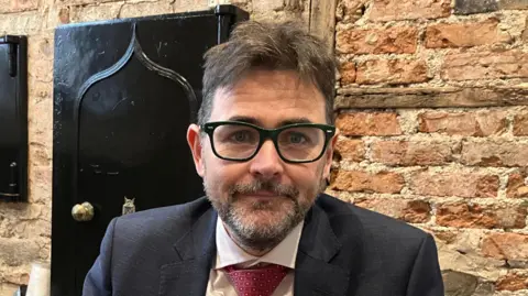 Cormac McDonnell photographed in his office (a brick walled building with files on the floor by his wooden desk). Cormac is wearing a navy suit with a white shirt and a red tie. He has short grey hair and is wearing black framed glasses.