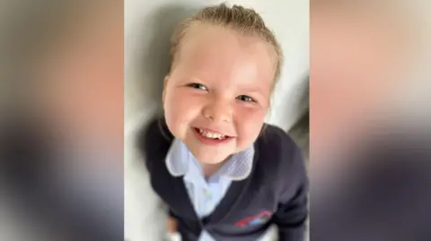 Arabella smiling at the camera. The image is taken from the top, looking down at the little girl. Her smiling face is in focus, with the rest of the image blurred. She has blonde hair tied back and blue eyes. She is wearing a blue and white blouse and blue blazer.