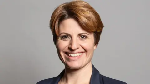 UK Parliament Official portrait of Emma Hardy, who has medium length brown hair and is standing in front of a neutral grey background.