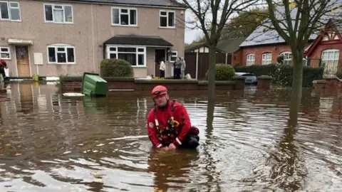 West Midlands Fire Service A man in red overalls standing waist deep in water in front of a row of houses