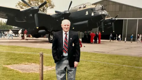 Family Handout Ivor Taylor standing in front of a Lancaster bomber, which is on display at an airfield. He is wearing grey trousers, a white shirt, black blazer and a red and black striped tie.