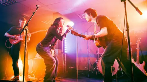 Ella Calver Young female and male lean into each other as they sing on a stage. The woman wears glasses and holds a microphone and the man plays a guitar. Another guitarist is playing on stage too.