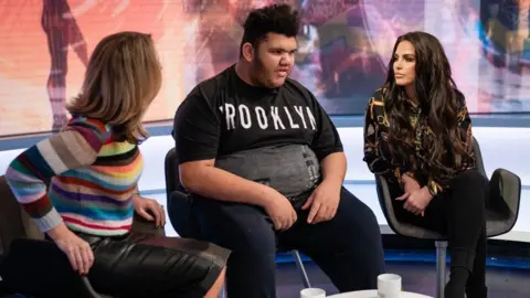 Katie and Harvey Price sitting on stools next to BBC presenter Victoria Derbyshire. They are engaged in conversation inside a TV studio.