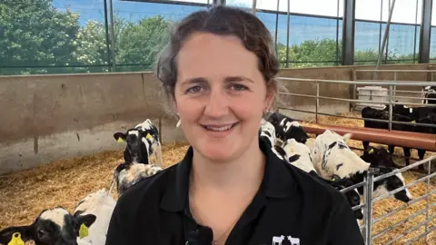 A woman with brown hair stands in front of about seven or eight cows in a barn.