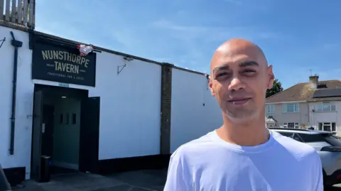 A man with an earring and a shaved head, wearing a white T-shirt. He smiles and stands outside his pub, the Nunsthorpe Tavern.