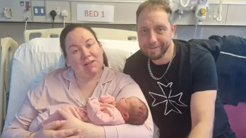 Emma Brown A man and woman sit together in a hospital bed holding a newborn baby in a pink baby grow.