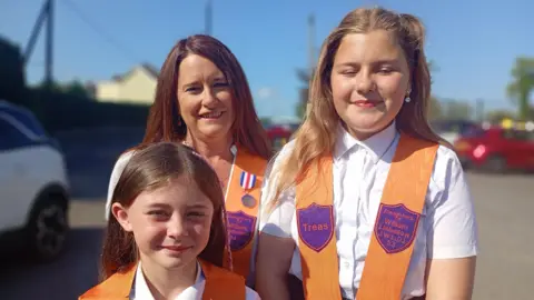 A woman is standing with two girls. All three are wearing bright orange sashes with purple badges. 