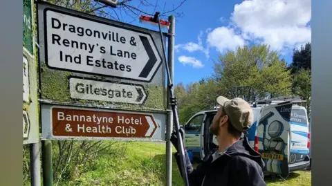Jonny Linden has a cap on in the sunshine and is cleaning a mossy road sign with a brush. The sign reads 'Dragonville & Renny's Lane Ind Estates and Gilesgate. Beneath is a brown sign reading 'Bannatyne Hotel & Health Club".