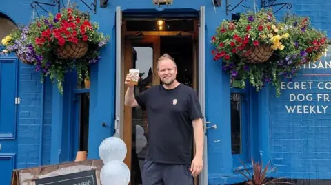 Cambridge Pub2Pub Tom Foddy, holding a glass of beer in his right hand, outside a pub. He is smiling. He is wearing a dark T-shirt and has short fair hair and a beard. The pub is painted blue behind him and has two large hanging baskets, either side of the doorway. 