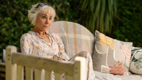 UKTV Zoë Wanamaker sat on a cushioned outdoor sofa with floral pillows to the right of her and a wooden outdoor chair blurred in the foreground. Behind are bushes.