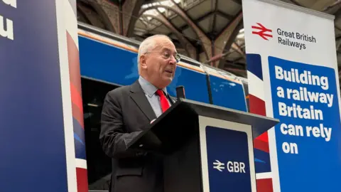 Zoie O'Brien/BBC Lord Hendy at Liverpool St Station, London. He is standing at a podium which has a logo saying GBR on it. He has grey hair and is wearing glasses, a grey suit, a checked shirt and red tie. Behind him is a railway carriage in the new blue, red and white livery. 