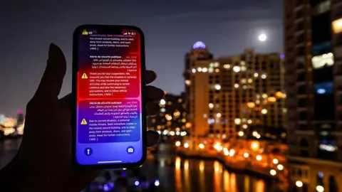 Phone held up to the camera at night, on it are a stream or emergency messages. In the background are a number of high-rise buildings