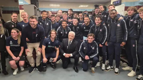 Scunthorpe United A man in a full black and white suit and tie with white shirt is knelt down on the floor surrounded by a team of mainly young male football players wearing a navy kit. They appear to be inside a locker room.