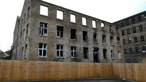 A mill building which has been damaged by fire, the roof has gone as have many of the windows and there is a fence around which looks new.