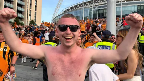 BBC / Linsey Smith A shirtless man wearing sunglasses has sunburned skin and a big smile as hestands in front of Wembley Stadium