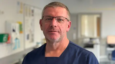 Dr Brian McKaig looks directly at the camera, he has grey hair and is wearing glasses and dark blue hospital scrubs