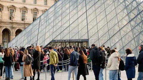 Imágenes de Getty de visitantes visitando el exterior de la entrada del Louvre, que tiene un patrón de pirámide de vidrio y un patrón entrecruzado negro.