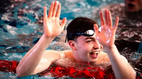 Euan Duff A close-up of Joel Watterson in the pool. He leans on a float in the water. He has dark hair and wears goggles. He is open mouthed and smiling with both hands in the air.