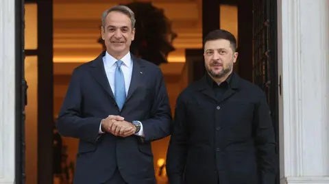 Anadolu via Getty Images A man in a blue suit and tie and white shirt smiles for the camera alongside another man dressed all in black.