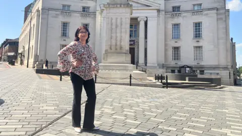 Amy Holmes/BBC A woman in a floral blouse poses in a paved square in front of a large stone building that is Luton Town Hall on a sunny day.