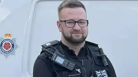 Lancashire Police Head and shoulders of Alex Rose in full police uniform standing at the side of a white police van with the Lancashire Constabulary crest to the left. He has short dark hair, a short beard and black-framed glasses