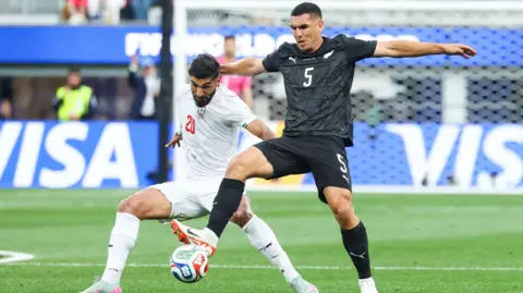 EPA/Shutterstock Match action between New Zealand and Iran. A New Zealand player shields the ball from his opponent.