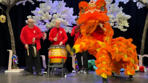 BBC A bright orange lion puppet is dancing to drums and cymbols. 