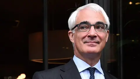 Getty Images Head and shoulder shot of a smiling man wearing dark-rimmed glasses, a dark grey suit jacket and a navy blue tie with white polka dots. He is standing against a dark backdrop.