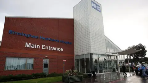 A photo of the outside of a hospital. It says Birmingham Heartlands Hospital Main entrance on red brick. Then there is a white post which says NHS in blue.