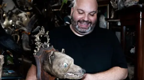 John Fairhall/BBC A man with a dark beard and wearing a black tshirt smiles at a taxidermy of an fox-like mammal'ss head mounted on a piece of wood, which he is holding 