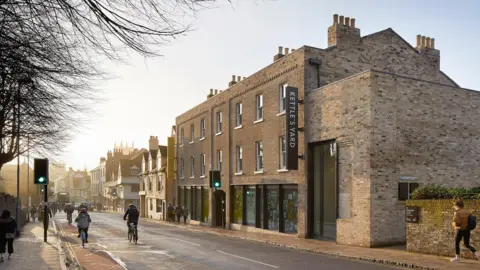 View of Kettle's Yard from the street outside. The sun is setting and people are walking and cycling past the gallery. It is a brick building with black windows and a large black sign sticking out the front.