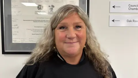 Martin Heath/BBC Kate Jolly with long white hair, wearing a black sweater and smiling at the camera. There is a white wall behind her with a certificate in a black frame.