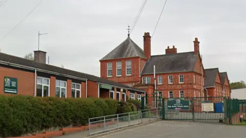 Google The closed green gates outside Earls Barton Primary School. It is a Victorian building, with a more modern modular building in the foreground. It is a cloud day.