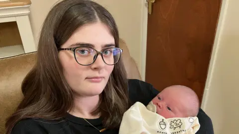 Aimee holding her baby. Aimee is wearing black top, glasses and gold necklace. She has long dark brown hair. The baby is wearing a cream. 