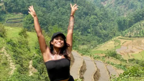Juliana Marins raising both hands in the air, in a black cap and black tube top. In the background are green paddy fields.