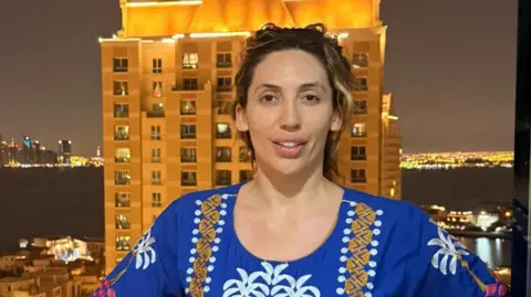 Katie Evans smiles as she poses in a tradiotianl embroidered Arabian dress on a balcony at night with high-rise building behind