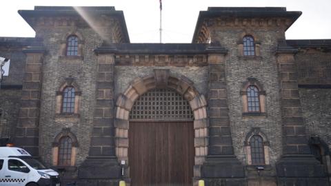 Main entrance of HMP Wandsworth showing the big oak door and a white police van parked outside on the left. The entrance is a large wooden arched doorway and there are stone mullioned arch windows on either side of the main door.