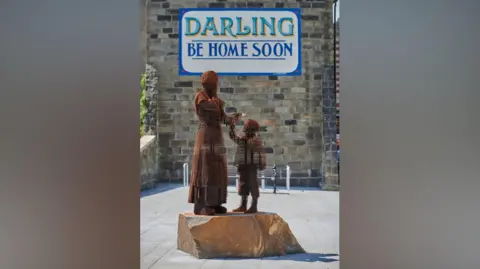LDRS The sculpture of a mother and son stands in front of a sign reads "Darling Be Home Soon".