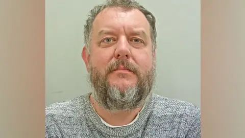 Lancashire Police Mugshot of Matthew Gilkes. He is wearing a grey marled sweater and has greying short curly hair and a beard.