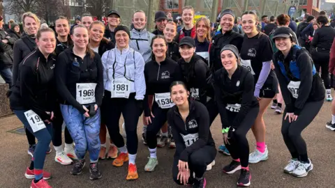 A group of 20 young women pose for the camera. Some are standing and others are crouching down. They are all wearing running trainers, leggings and long sleeve tops and have numbers pinned onto their tops. 