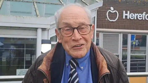 LDRS Councillor Matthew Engel, a bald man who is wearing glasses and a blue shirt with a black and white striped tie and a brown coat. He is standing outside a council building with glass entrance and railings.