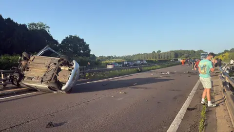 A silver car is pictured on its roof and up against the central reservation of the motorway. A man in a blue t-shirt and beige shorts is standing on the side of the motorway on the phone. Several other people are pictured in the background standing on the motorway. Debris is scattered across the road.
