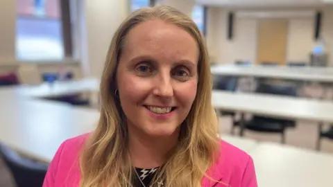 A head and shoulders photograph of councillor Naomi Tweddle. She is standing in a room with table and chairs behind her and is wearing a bright pink suit jacket.