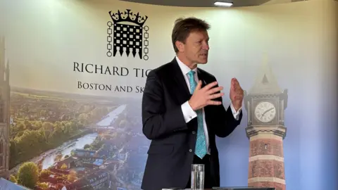 MP Richard Tice standing in front of a display board showing an aerial view of the river in Boston. He is wearing a suit and a blue tie.