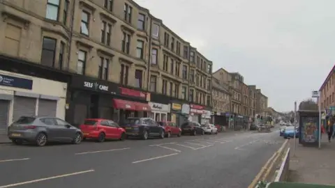 BBC Neilston Road, Paisley,  street with cars parked on either side, image taken from the pavement near a bus stop with tenement flats opposite and shops on the ground floor