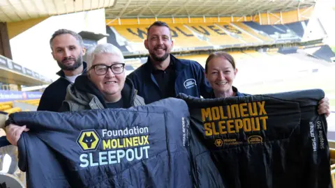 Four people are standing inside a football stadium. Two men are standing at the back, wearing jackets that read "Good Shepherd". Two women are in front holding sleeping bags that read "Molineux sleepout". There are thousands of football seats in stands behind them in a gold colour. The word "Wolves" is spelt out in black on them. 