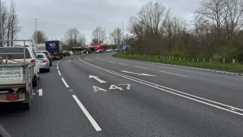 Siva of Elm Queuing traffic on the right, heading towards a roundabout. The lanes on the right hand side are empty. In the far distance is traffic queuing as it goes around the roundabout and beyond that a large bright red lorry. 
